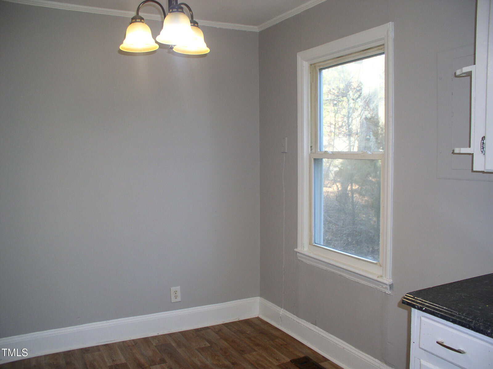 A simple, empty room with light gray walls, a single window, wooden floor, white trim, a ceiling light fixture with three lamps, and a partial view of a countertop and cabinets on the right.