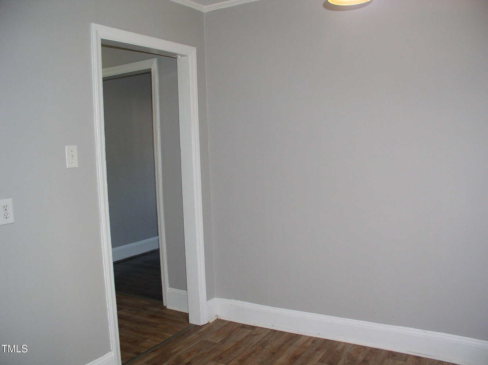 A small, empty room with light gray walls, white trim, wood-look flooring, and an open doorway leading to another room with similar decor. A ceiling light is partially visible at the top right.