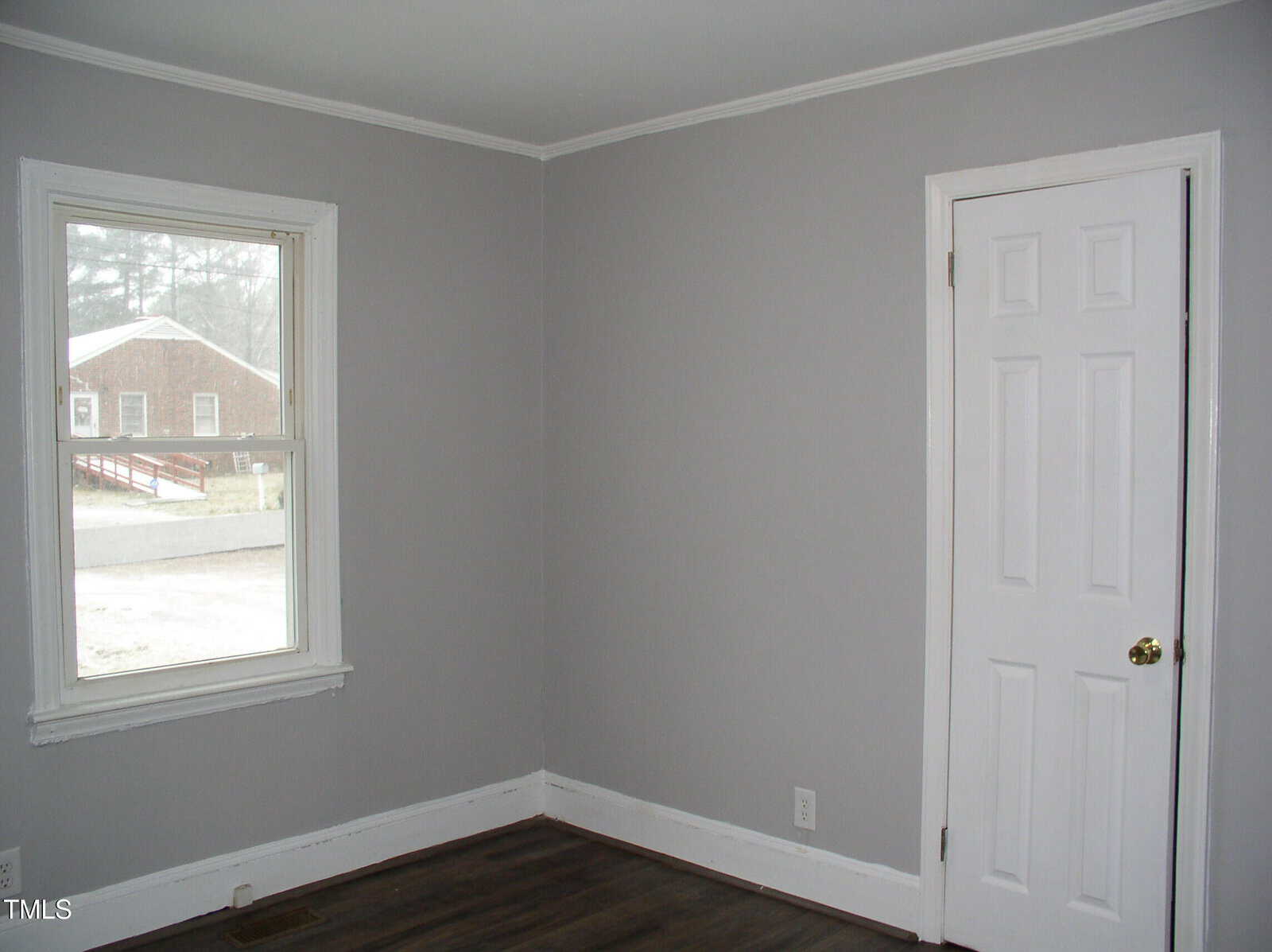 A small, empty room with light gray walls, white trim, a white door with a gold knob, and a window letting in daylight. The floor is dark wood, and there is an electrical outlet on the wall.