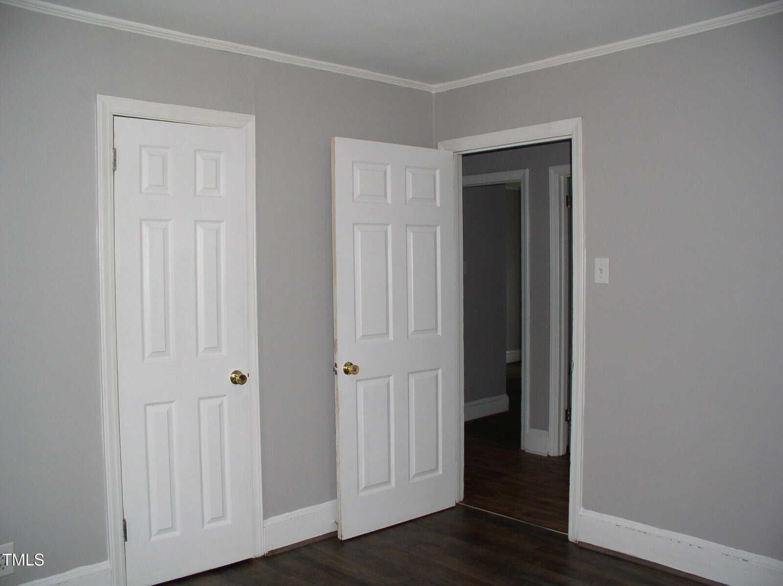A room with light gray walls, dark wood flooring, and white trim. There are two white doors, one closed and one open, showing an adjoining room with similar decor.