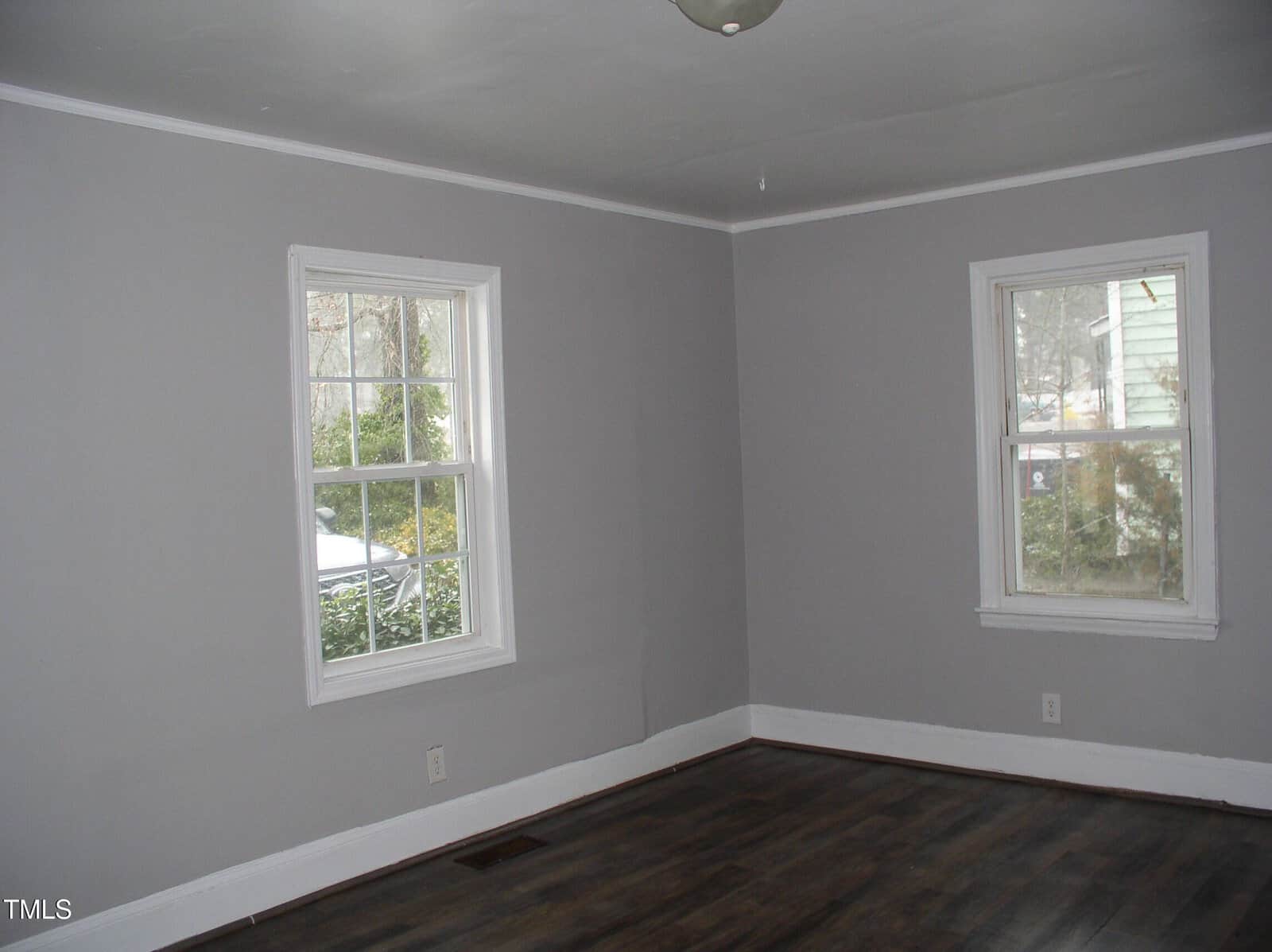 Empty room with gray walls, white trim, dark wood floors, and two windows letting in natural light.