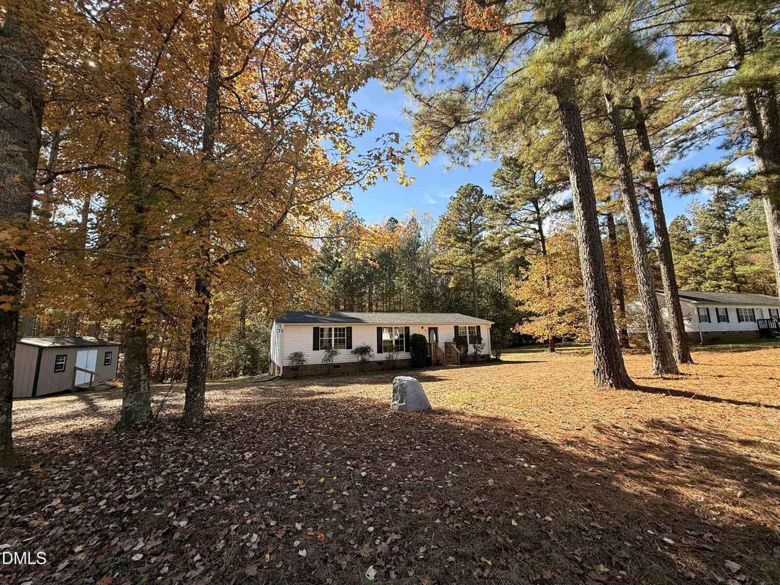 A single-story white house with a front porch sits among tall pine and deciduous trees with autumn leaves, under a clear blue sky. A large rock and scattered leaves cover the front yard.