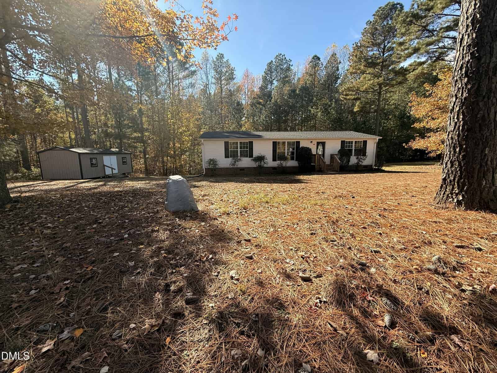 A white single-story house with dark shutters sits in a spacious yard surrounded by tall pine trees and autumn leaves. A small shed is to the left, and sunlight filters through the trees on a clear day.
