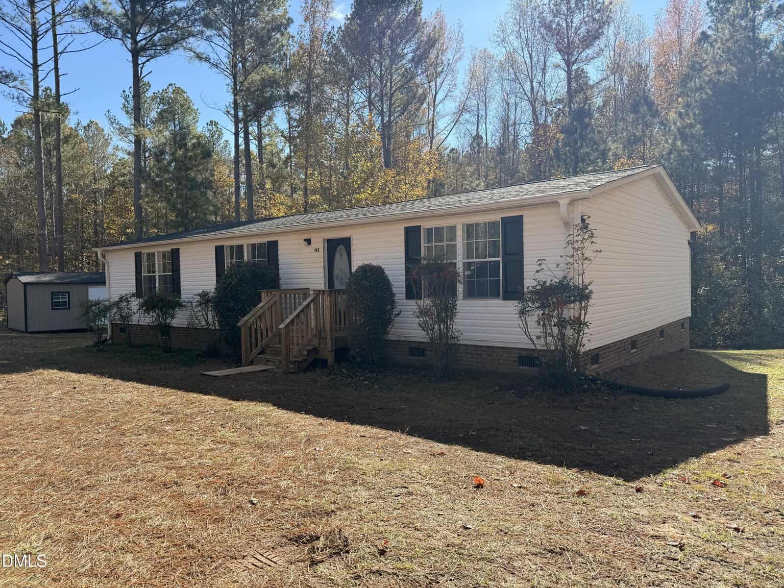 A white, single-story manufactured home with black shutters and a small front porch sits on a grassy lot surrounded by tall trees under a clear sky. A gray shed is visible in the background to the left.