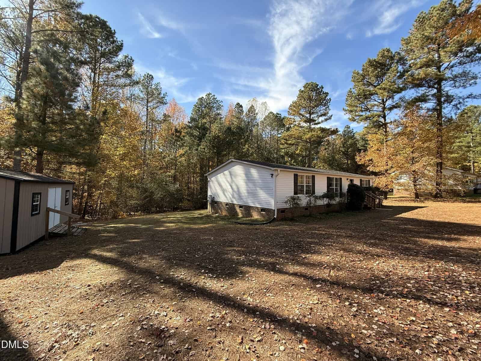 A white, single-story house with a front porch sits in a spacious, sunny yard surrounded by tall pine trees with autumn foliage. A small shed is visible to the left of the house.
