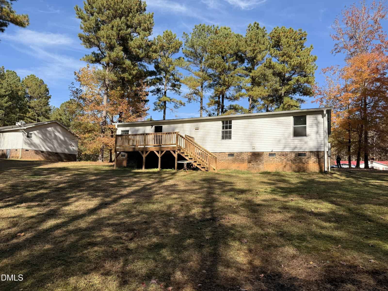 A single-story house with a brick foundation and white siding, featuring a wooden deck and stairs, sits on a grassy yard surrounded by tall trees with autumn foliage under a blue sky.