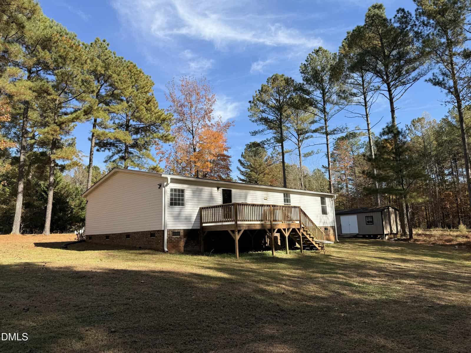 A white, single-story house with a wooden deck sits in a spacious yard surrounded by tall pine trees under a blue sky with light clouds. A small shed is nearby. Shadows from the trees fall across the grass.