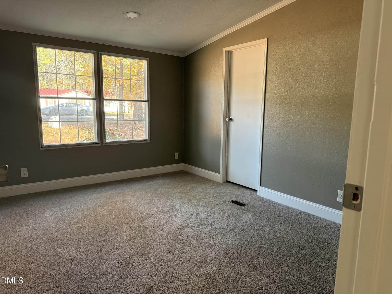 Empty carpeted room with gray walls, large window showing autumn trees outside, white baseboards, and a closed white door on the right wall. Natural light fills the space.