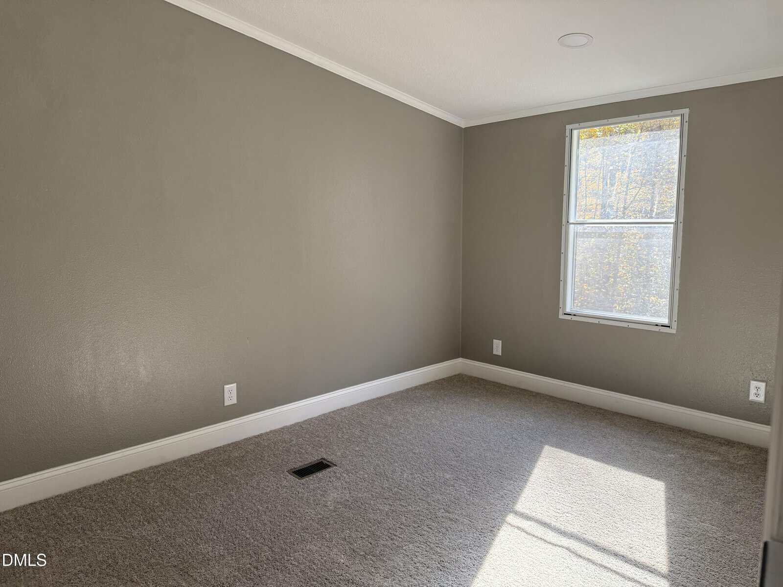 An empty room with beige carpet, gray walls, white baseboards, and a single window letting in sunlight. There are electrical outlets and a floor vent visible.