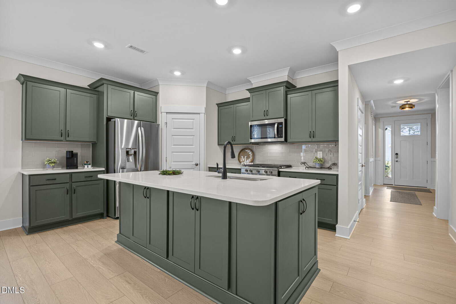 Modern kitchen with sage green cabinets, white countertops, stainless steel appliances, and light wood floors. A central island features a black faucet. A hallway with light walls and wood floors leads to the front door.