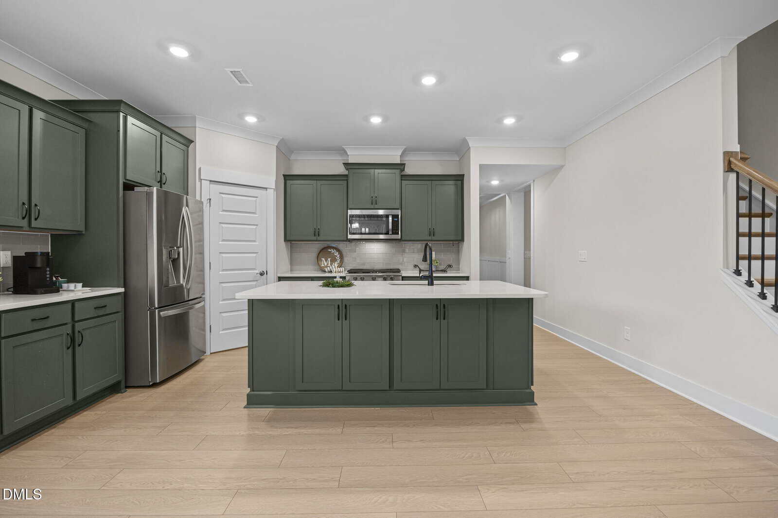 Modern kitchen with green cabinets, stainless steel appliances, a white island countertop, light wood flooring, and recessed ceiling lights. There is a staircase to the right and a white door to the left of the refrigerator.