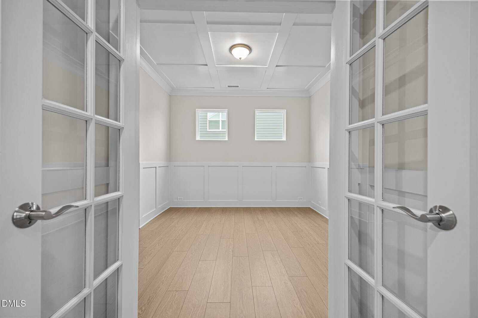 A bright, empty room with light wood floors, white paneled walls, a coffered ceiling, and two small windows, viewed through open double glass doors.