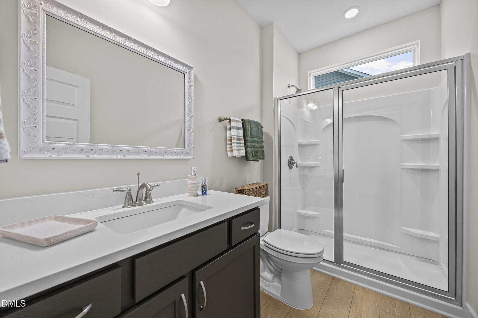 Modern bathroom with a white sink and countertop, dark cabinets, a large mirror, a toilet, and a glass-enclosed shower. White walls, wood-look flooring, and a window providing natural light. Towels hang by the shower.