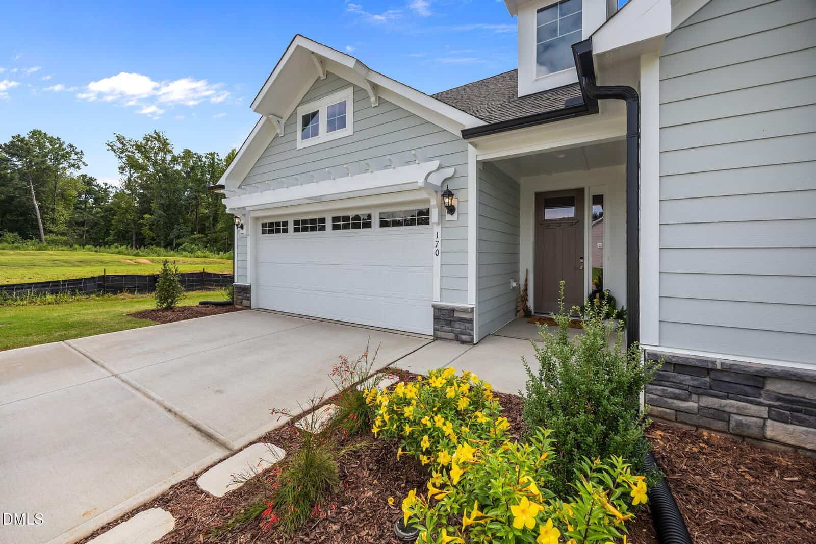 A modern light gray house with white trim and a double garage door, a concrete driveway, and a landscaped front yard featuring yellow flowers and green shrubs under a blue sky.
