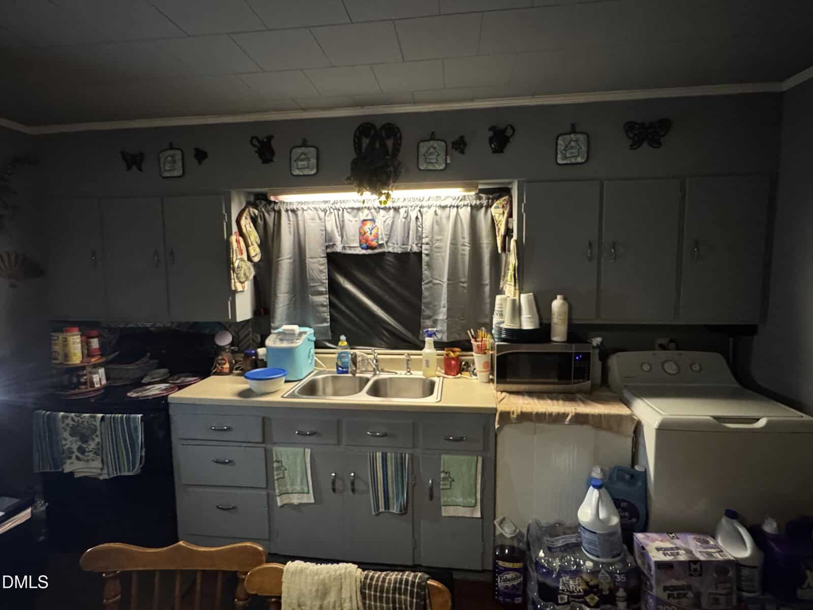 A dimly lit kitchen with gray cabinets, a double sink, a microwave, and a washing machine. Various household items clutter the counters, and decorative plates and butterflies line the wall above the window.