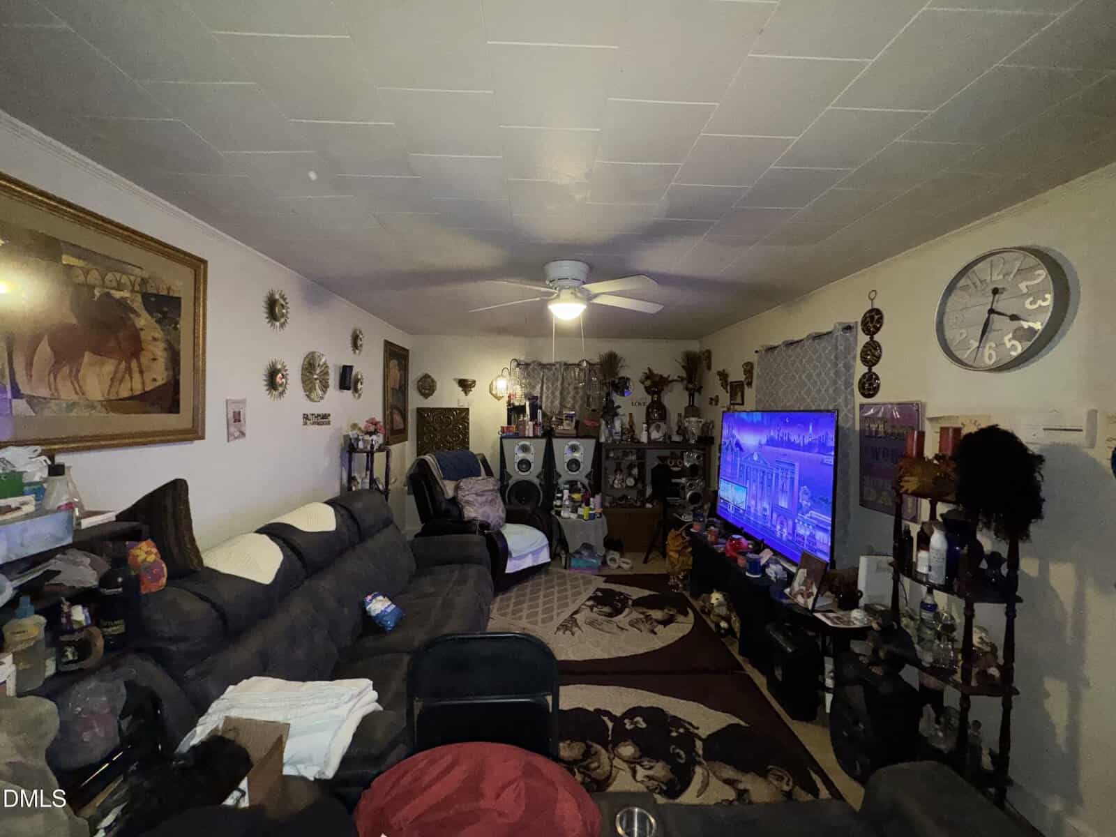 A cozy living room with dark sofas, a large TV displaying a cityscape, a ceiling fan, wall clock, framed art, shelves with decor, and various personal items. Neutral walls and dim lighting complete the space.