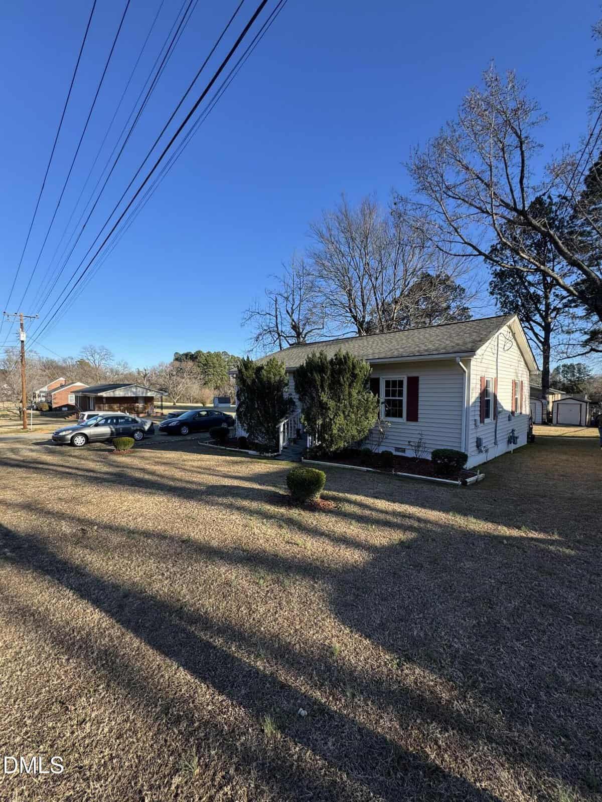Single-story white house with red shutters, small bushes in the front yard, several cars parked along the driveway, and tall trees without leaves under a clear blue sky. Power lines stretch overhead.