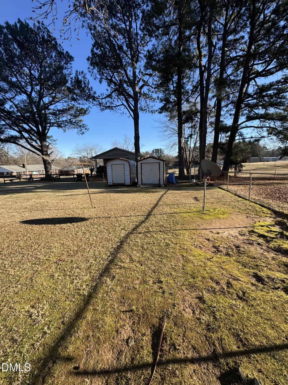 A backyard with patchy grass, a shed and garage in the background, surrounded by tall pine trees. A satellite dish and pole stand in the yard, with clear blue sky overhead.