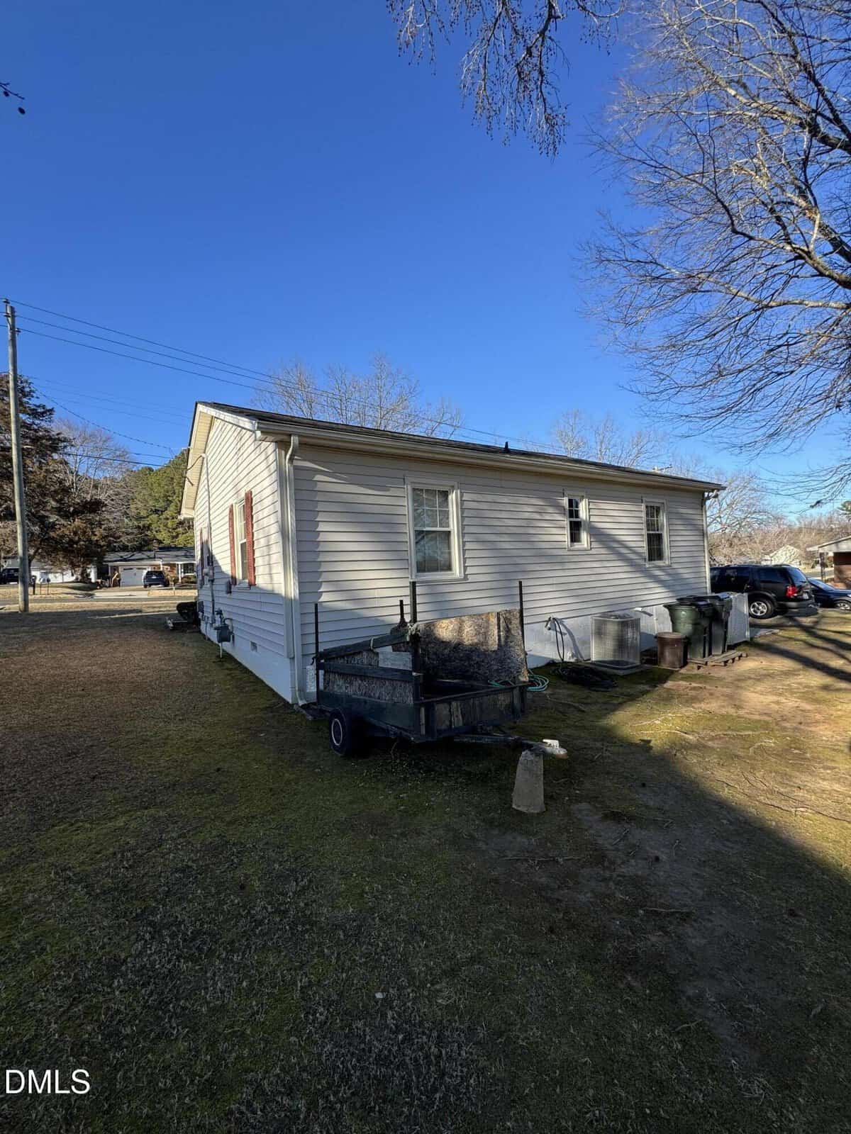 A small, single-story beige house with red shutters sits in a yard. A utility trailer and various items are beside the house. Leafless trees and a clear blue sky are in the background.