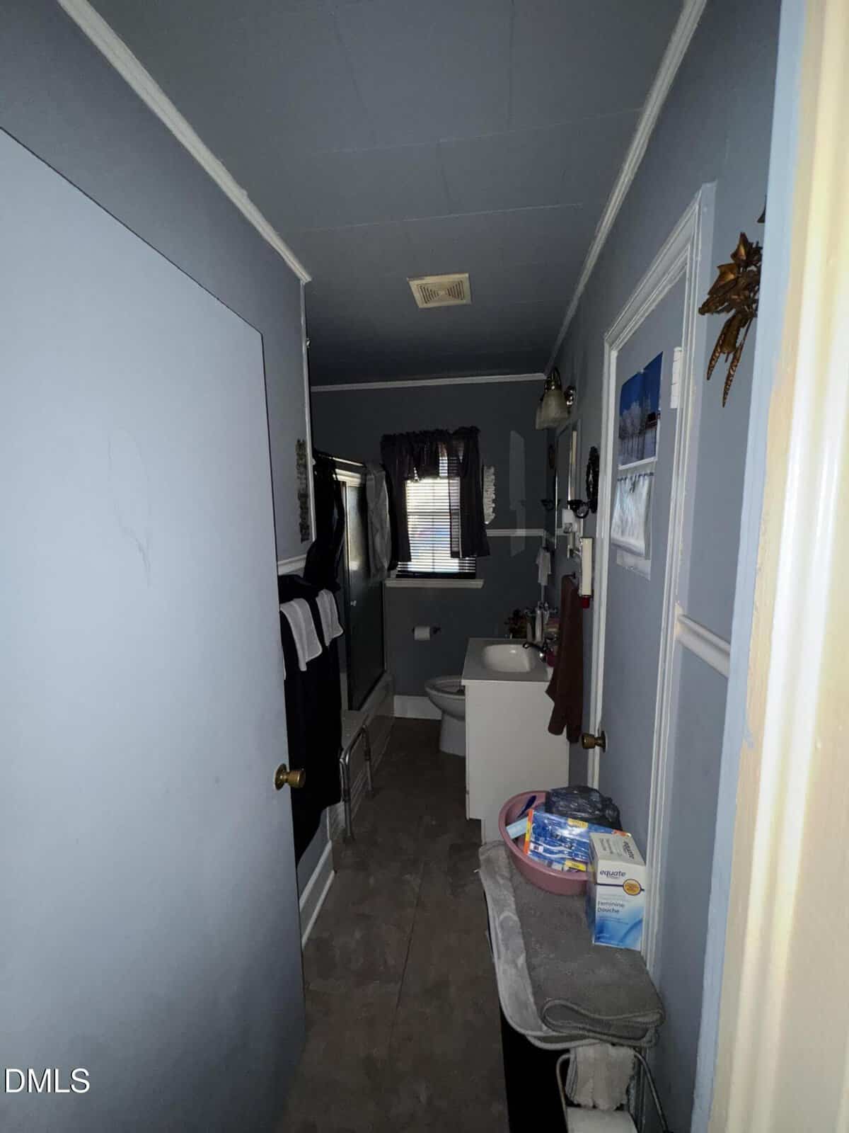 A narrow bathroom with gray walls, a white toilet, a sink, a bathtub with a black-and-white shower curtain, and a window. Shelves and towels hang on the walls, and toiletries sit on a small table by the door.