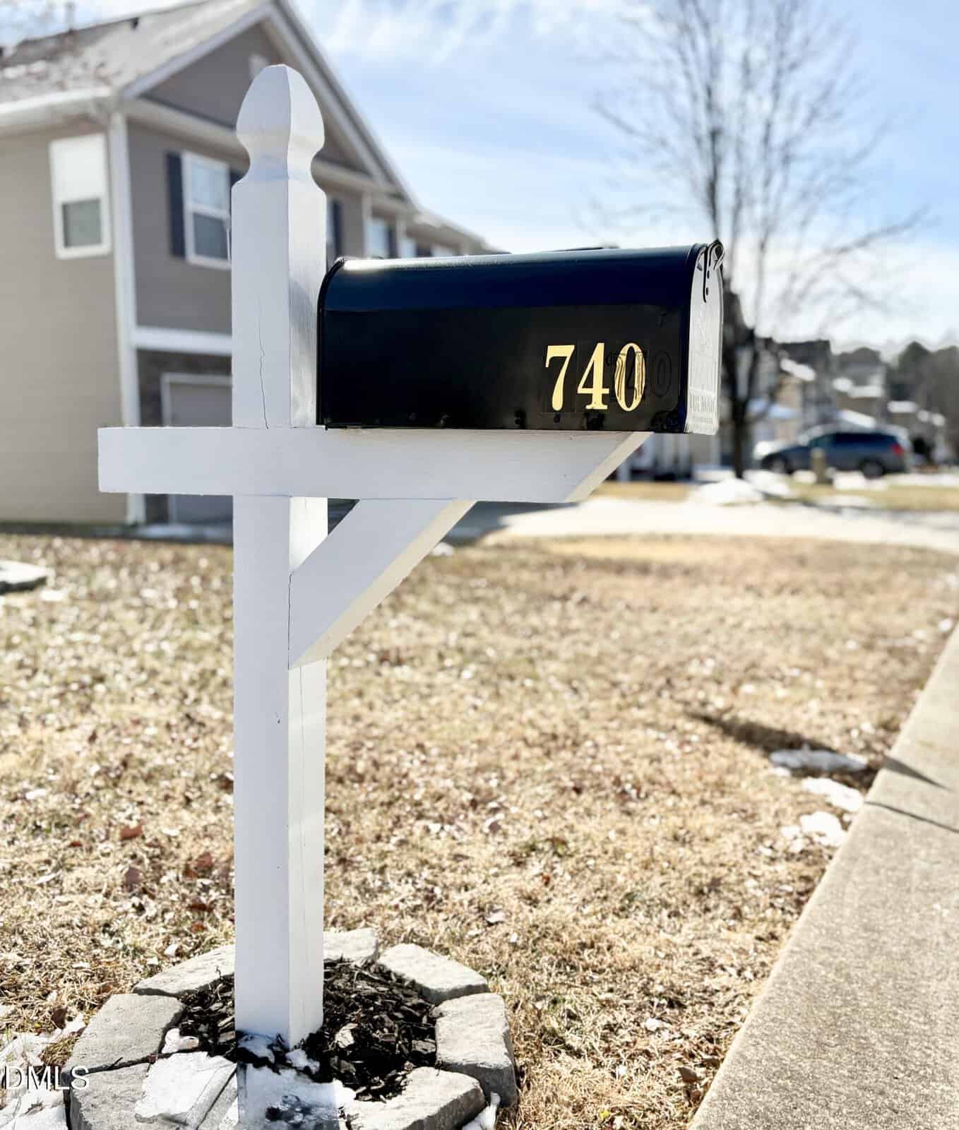 A black mailbox with the number 740 stands on a white post by a suburban street, with houses and patches of snow visible in the background on a sunny day.