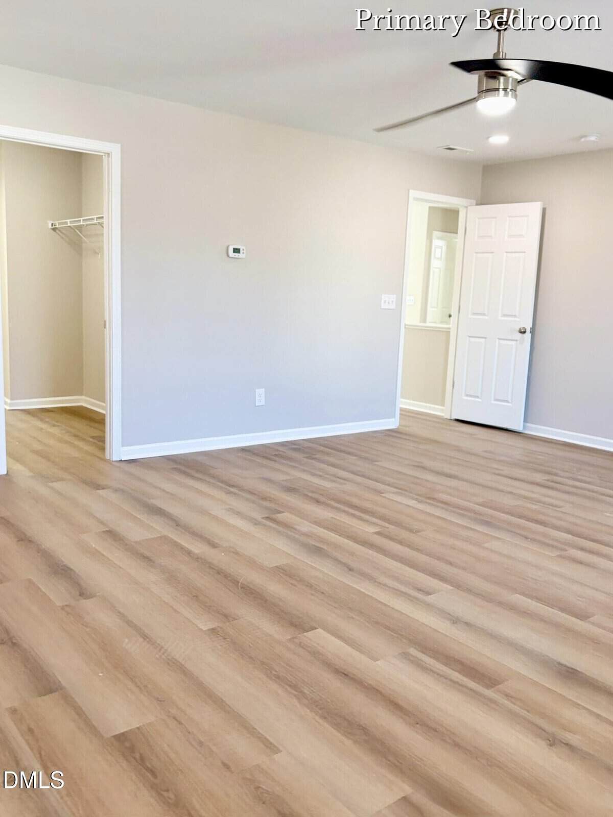 Bright, empty primary bedroom with light wood flooring, beige walls, a ceiling fan, open closet, and white trim. An open door leads to a hallway, and there is a small recessed area with a mirror.
