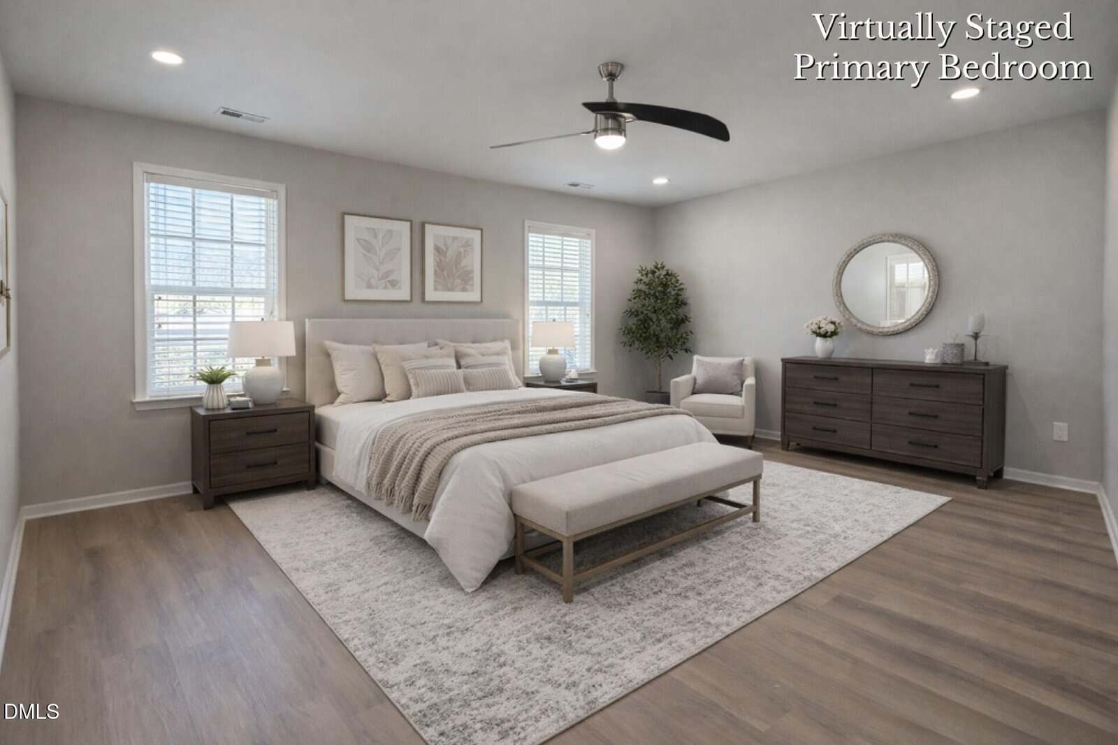 A spacious, bright primary bedroom with a large bed, two nightstands, a bench, dresser, armchair, potted tree, and decor. Light walls, wood flooring, and two windows provide a clean, modern look. Text reads "Virtually Staged Primary Bedroom.