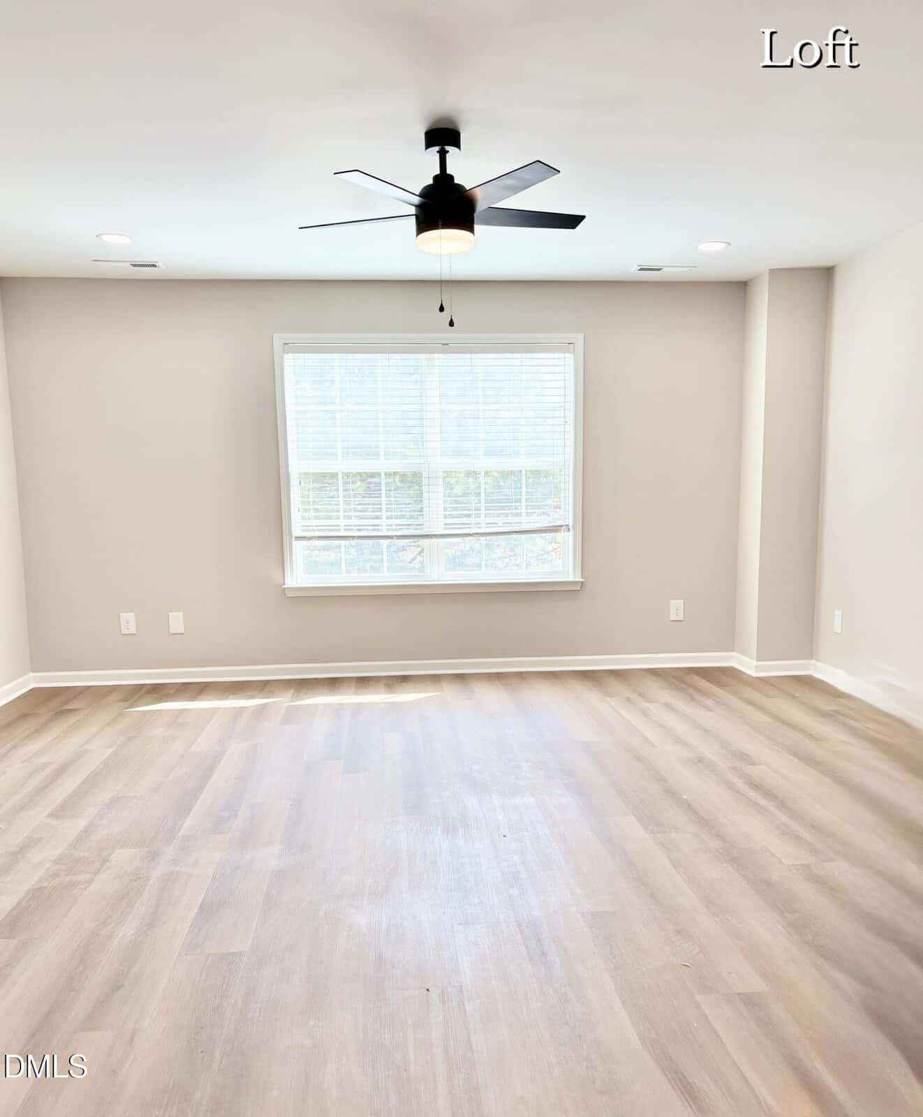 A bright, empty loft with light wood flooring, beige walls, a large window with white blinds, a modern black ceiling fan, and sunlight streaming in. The word "Loft" appears in the top right corner.