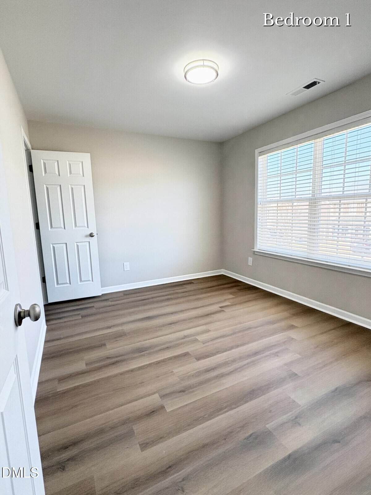 Empty bedroom with light gray walls, a white door, large window with blinds, and wood-look flooring. The room is well-lit with natural light. "Bedroom 1" is labeled in the top right corner.