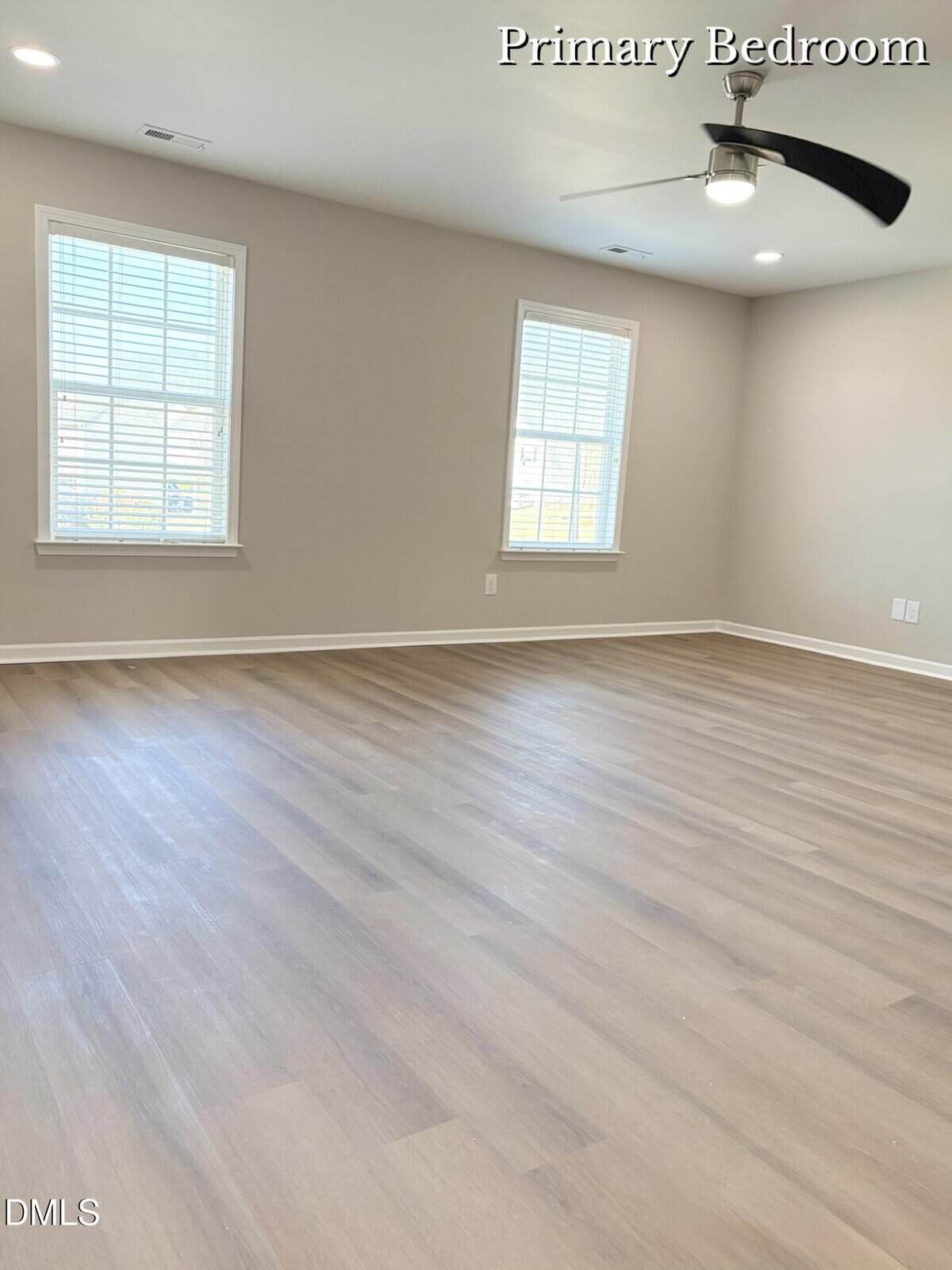 A spacious, empty primary bedroom with light-colored hardwood floors, two large windows with white blinds, beige walls, a ceiling fan with a light, and ample natural light.