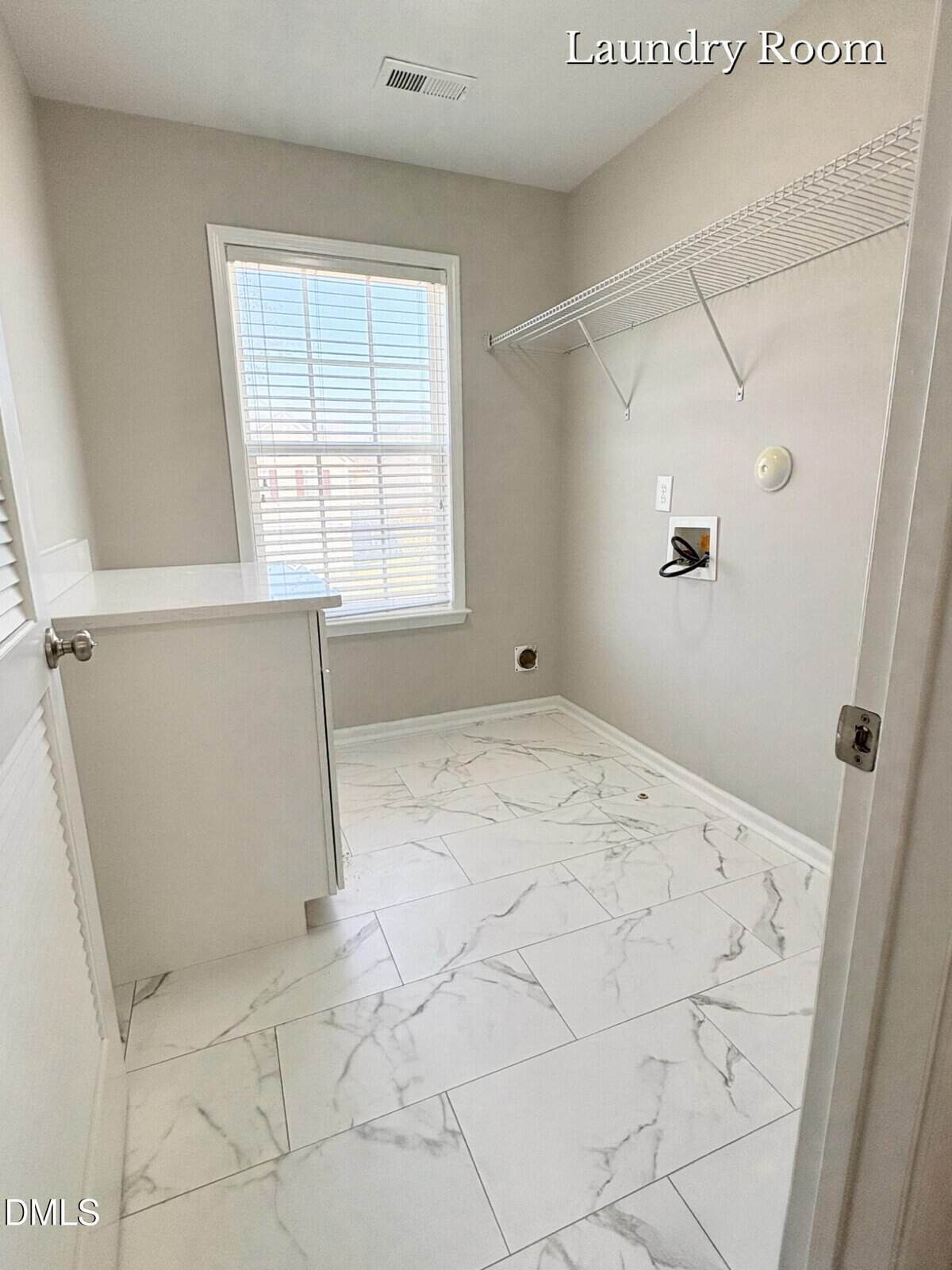 A bright, empty laundry room with white marble-patterned floor tiles, light gray walls, a large window, a white cabinet, and a wall-mounted wire shelf. "Laundry Room" is labeled in the upper right corner.