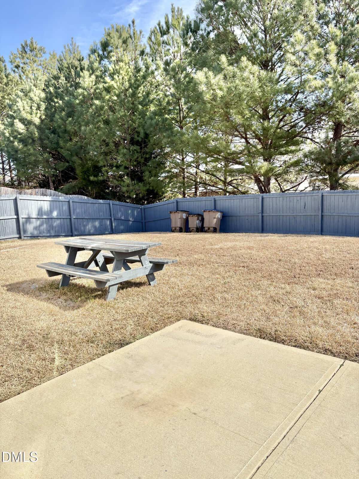 A backyard with dry grass, a small gray picnic table, three trash bins by a blue wooden fence, and a concrete patio in the foreground. Tall pine trees line the fenced perimeter.