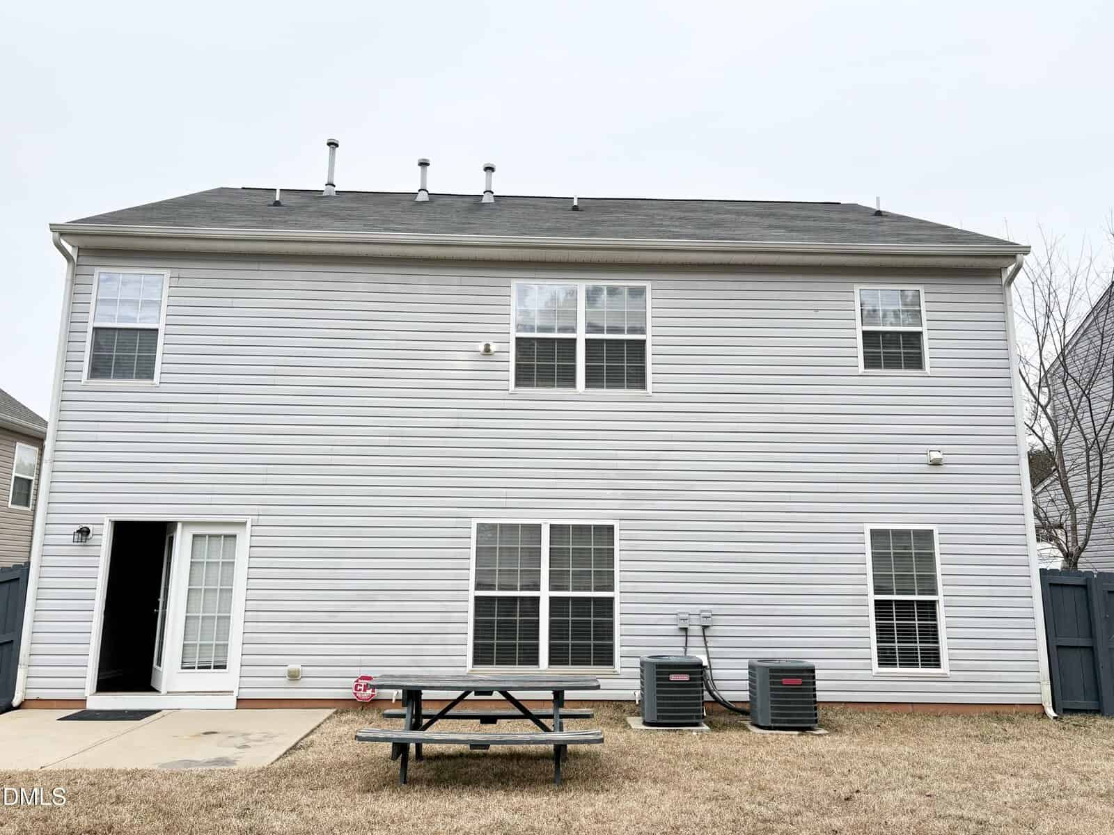 Back view of a two-story gray house with white trim, four windows, a glass door, two HVAC units, a picnic table on dry grass, and a fenced yard under a cloudy sky.