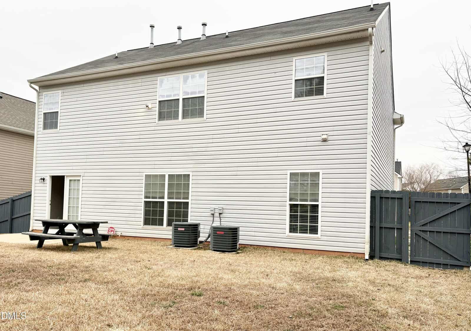 Two-story beige house with white trim, three visible windows, a fenced backyard, brown grass, two HVAC units, and a black picnic table. The sky is overcast.