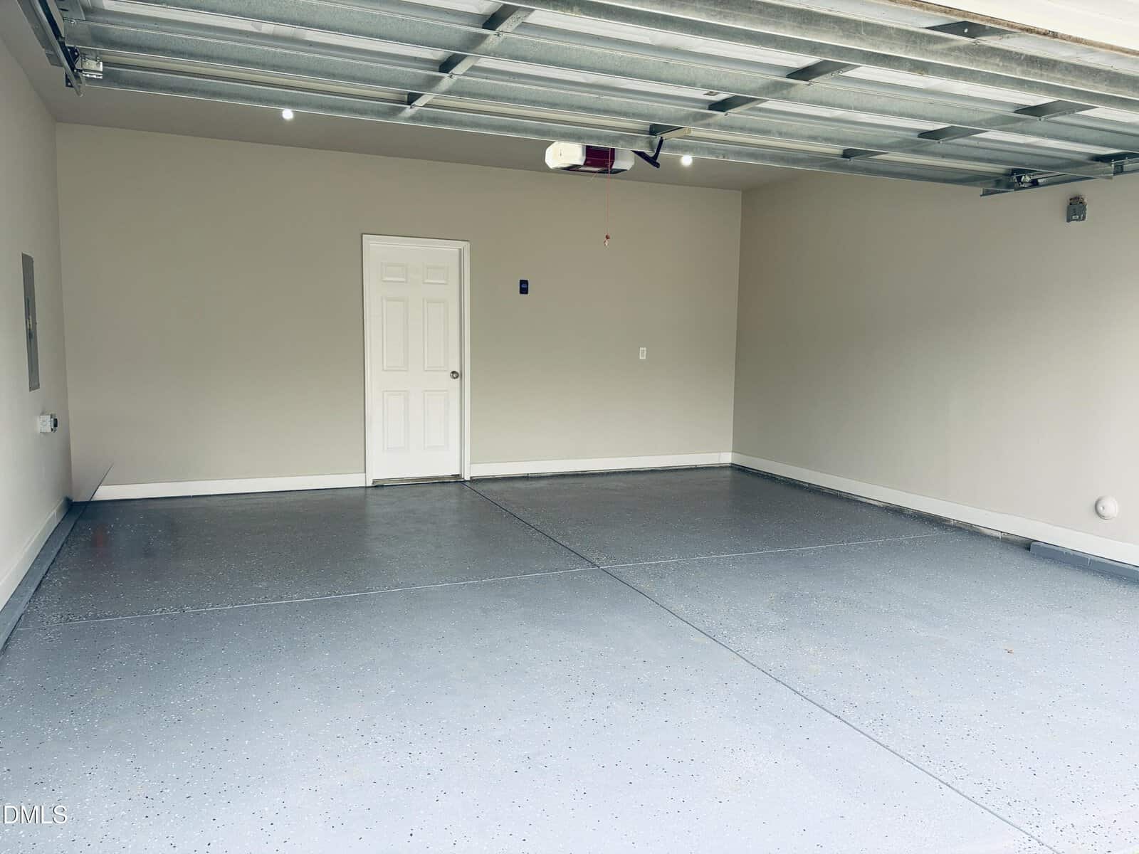 Spacious, empty two-car garage with light gray speckled floor, beige walls, and a white door at the back. The ceiling features a garage door opener and exposed tracks.