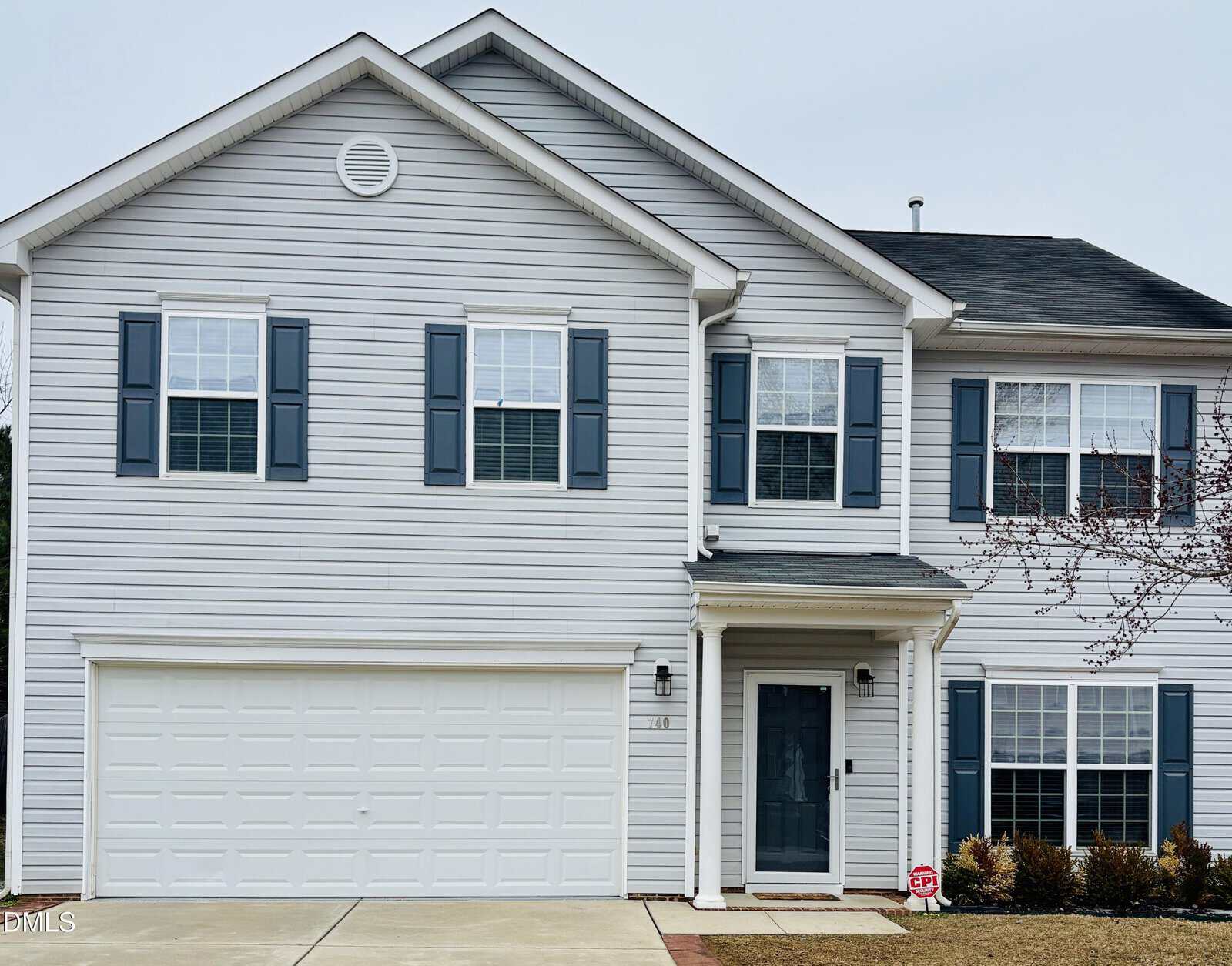 Two-story suburban house with light gray siding, blue shutters, and a double garage. There are several windows on both floors, a small front porch with a black door, and a neatly trimmed lawn with small shrubs.