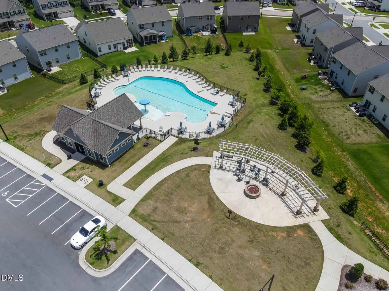 Aerial view of a residential community showing a large outdoor pool with lounge chairs, a clubhouse, a circular seating area with a pergola, and parked cars next to houses.