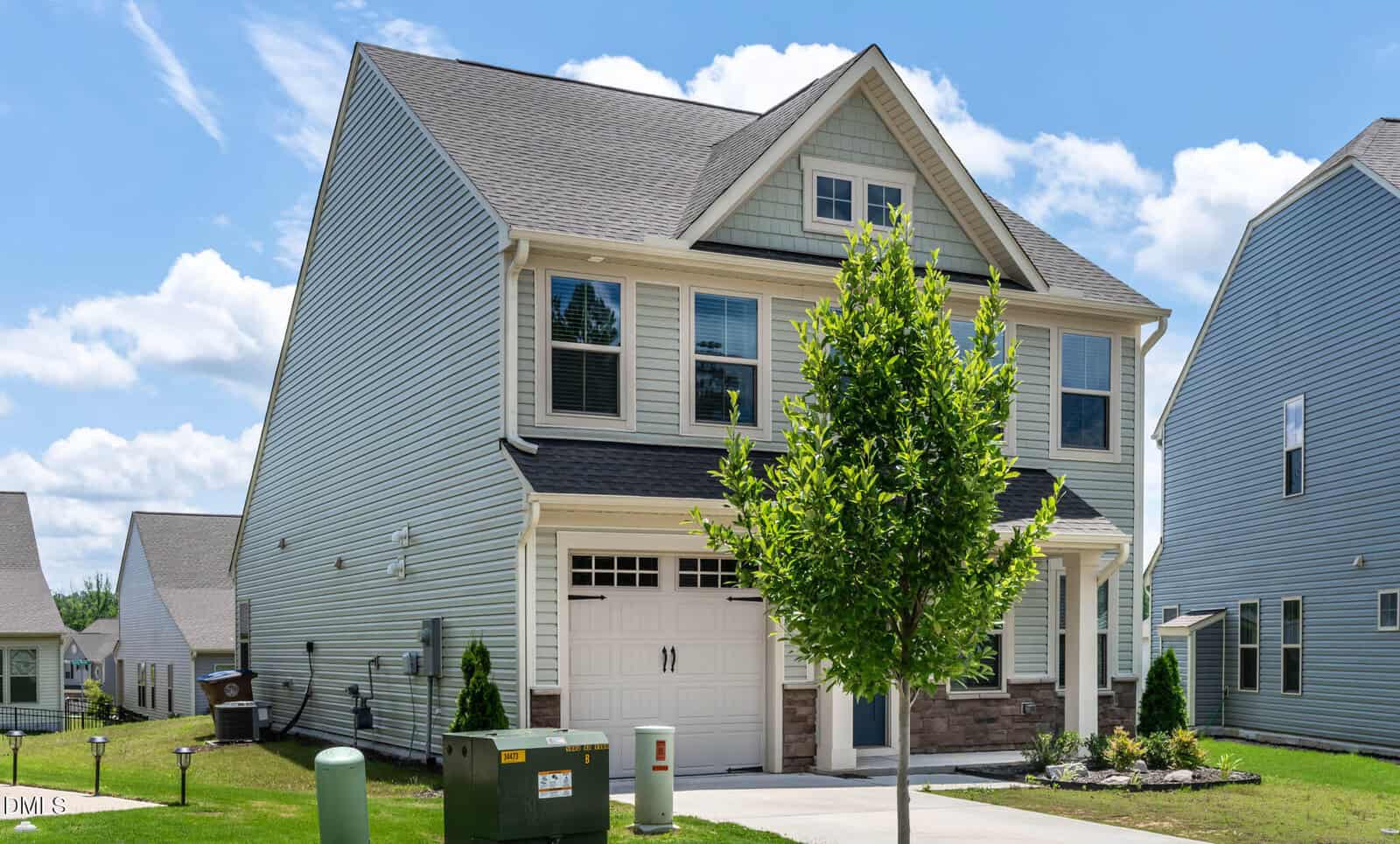 Two-story suburban house with gray siding, stone accents, and a front-facing garage. A small tree is planted in the front yard, with a driveway and green utility boxes visible near the street under a partly cloudy sky.