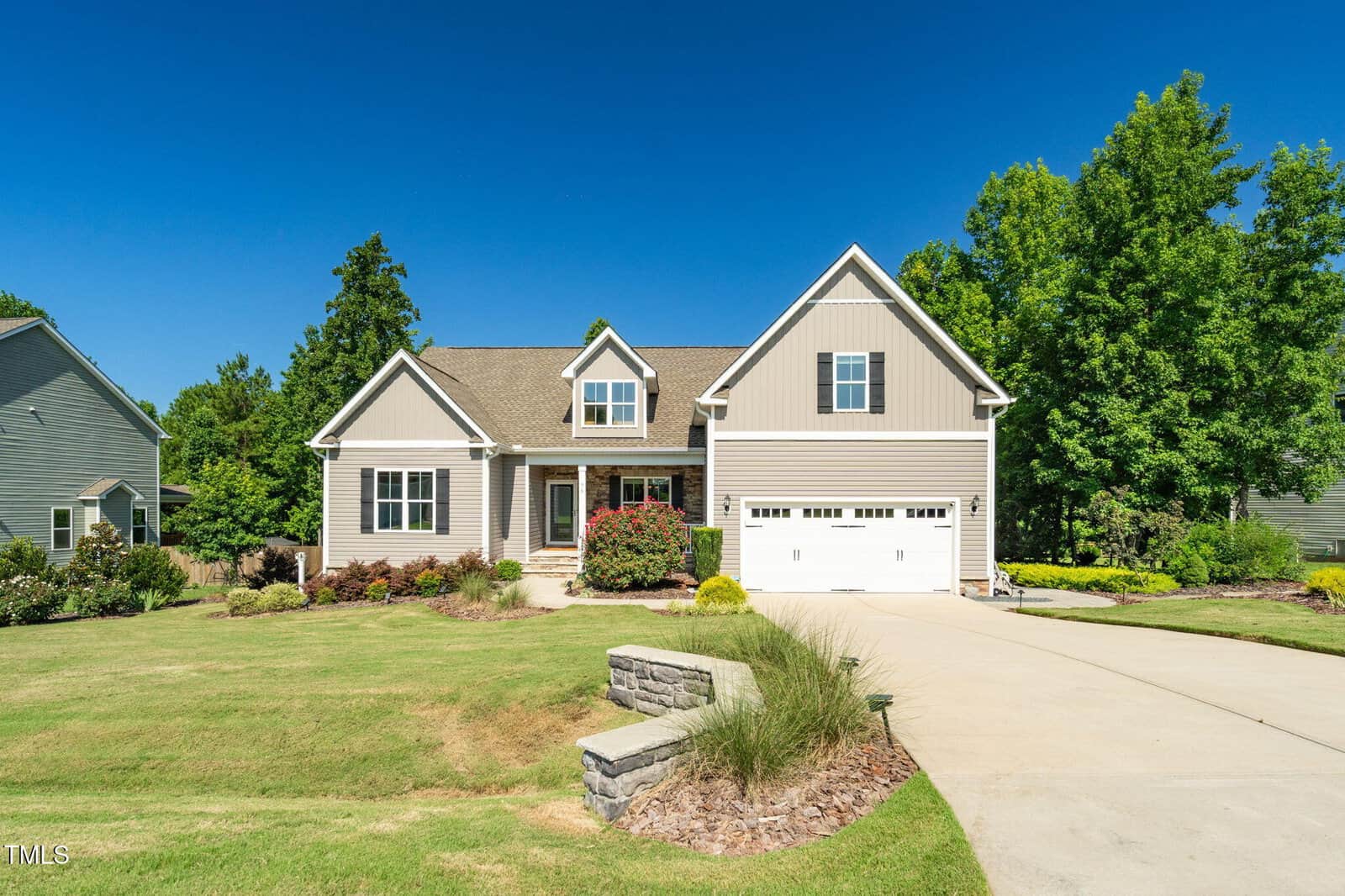 A beige two-story suburban house with white trim and a double garage sits on a well-maintained lawn with landscaping and a concrete driveway under a clear blue sky.