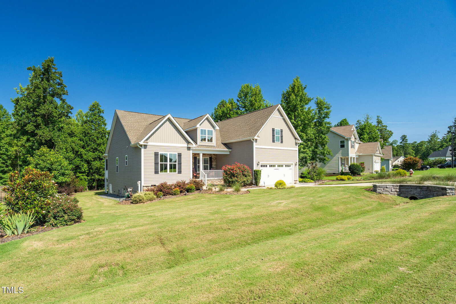 A single-family house with light-colored siding, a covered front porch, and an attached two-car garage sits on a large, well-kept lawn with landscaping. Other houses and trees are visible in the background under a clear blue sky.