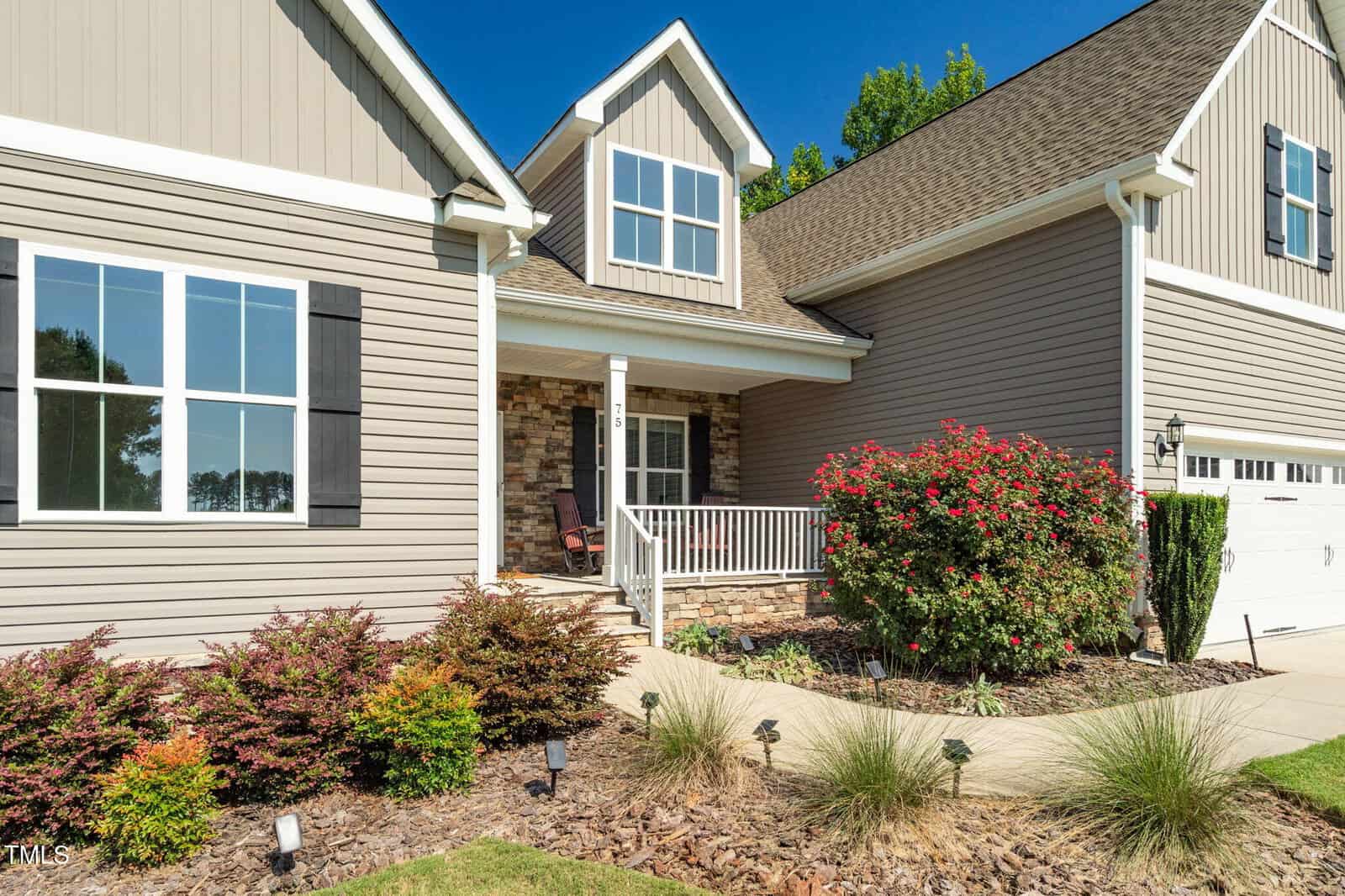 Single-story house with gray siding, white trim, and a gabled roof. The front porch has a white railing and two chairs. There are shrubs, mulch, and a flowering bush in the landscaped front yard.