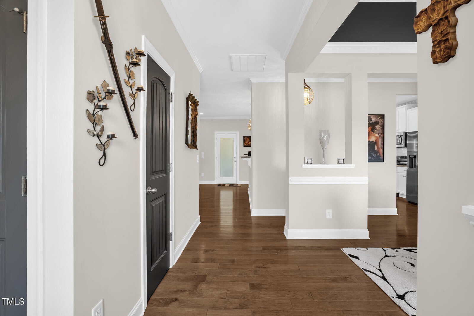 A hallway with light beige walls, dark wood doors, and hardwood floors leads to multiple rooms. Metal wall decor and a patterned rug are visible, with a glimpse of a kitchen on the right.