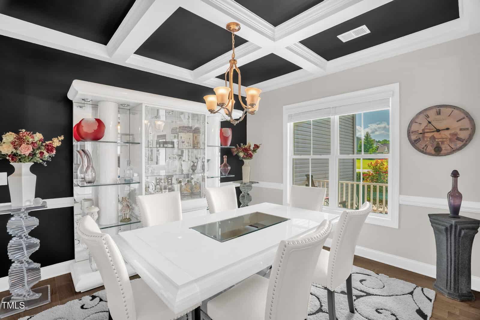 A modern dining room with a white table and six white chairs, a glass display cabinet, decorative flowers, a large wall clock, chandelier lighting, and a window with a view of greenery outside.