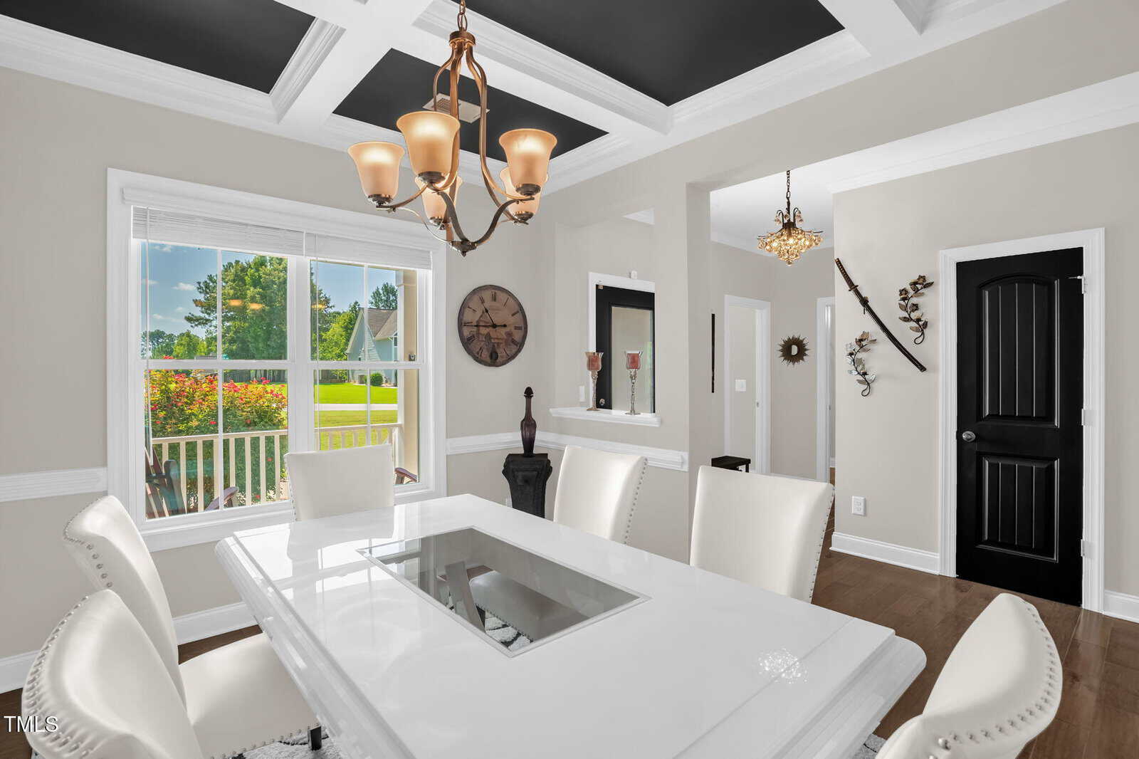 A bright dining room with white chairs and a white table, a coffered ceiling with black accents, chandelier, wall clock, decorative items, and a window showing a green lawn and trees outside.