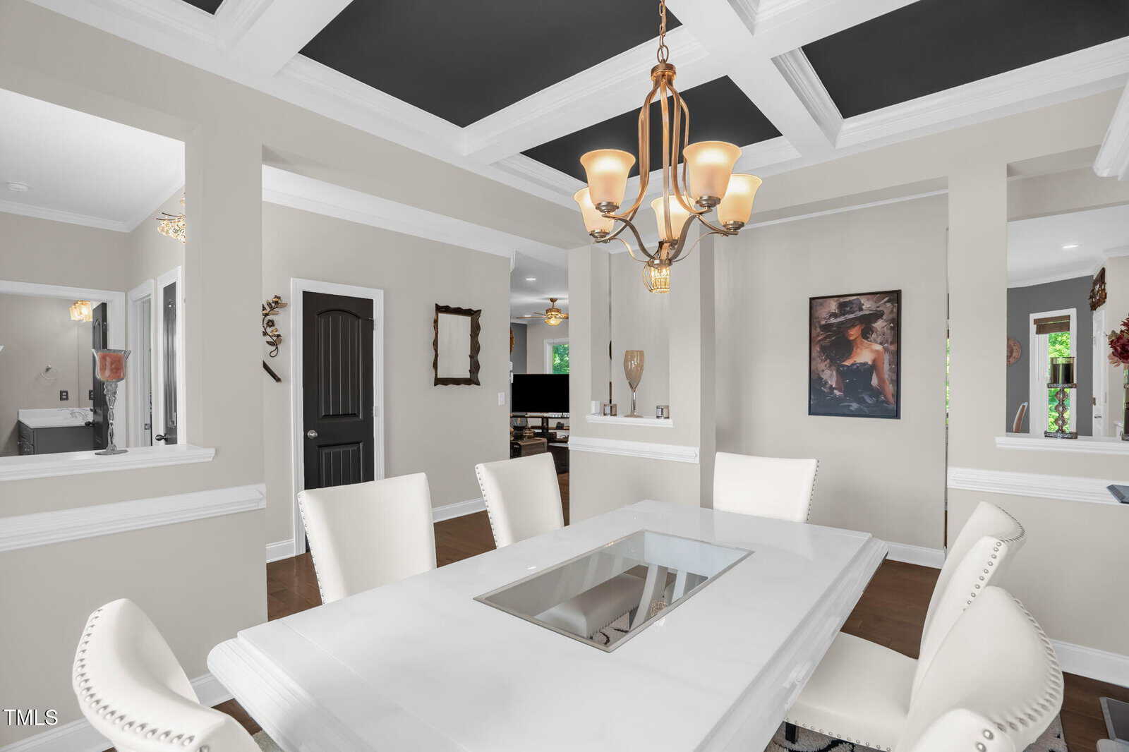 A dining room with a white table, six white upholstered chairs, a bronze chandelier, gray walls, white trim, coffered ceiling with black insets, and decorative art on the wall. There are open doorways and a view to another room.