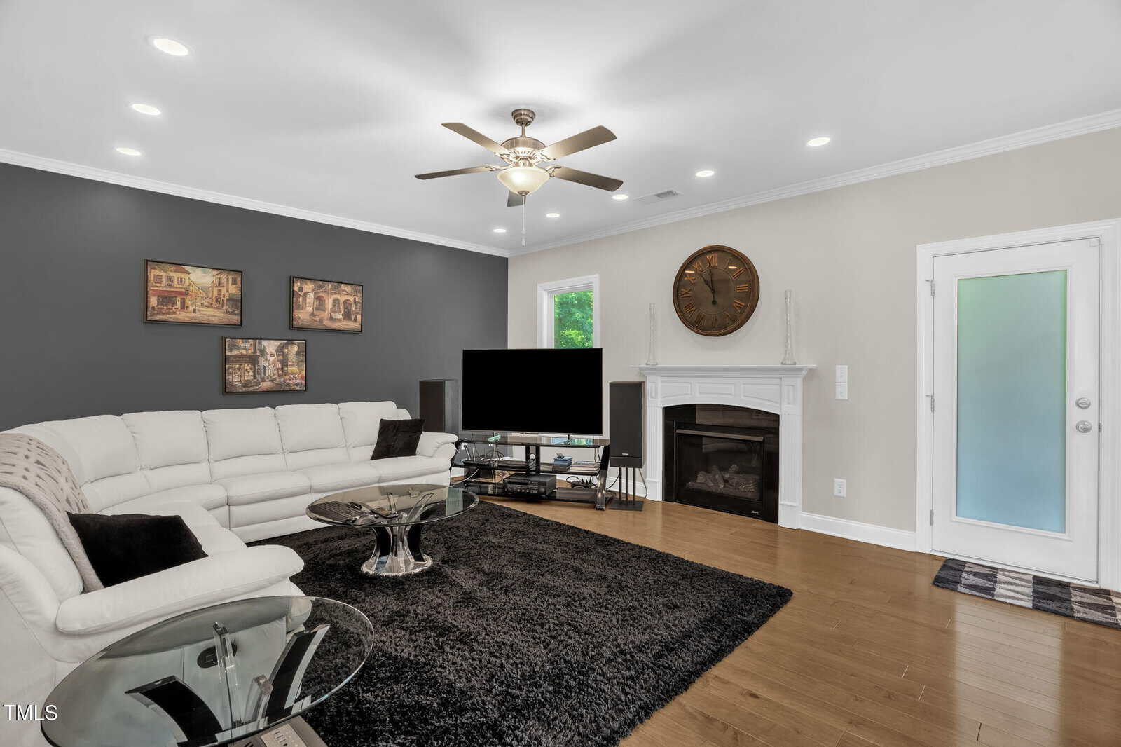 Modern living room with a white sectional sofa, glass tables, black rug, wall-mounted TV, fireplace with a large clock above it, and artwork on a gray accent wall. Ceiling fan and recessed lighting overhead.
