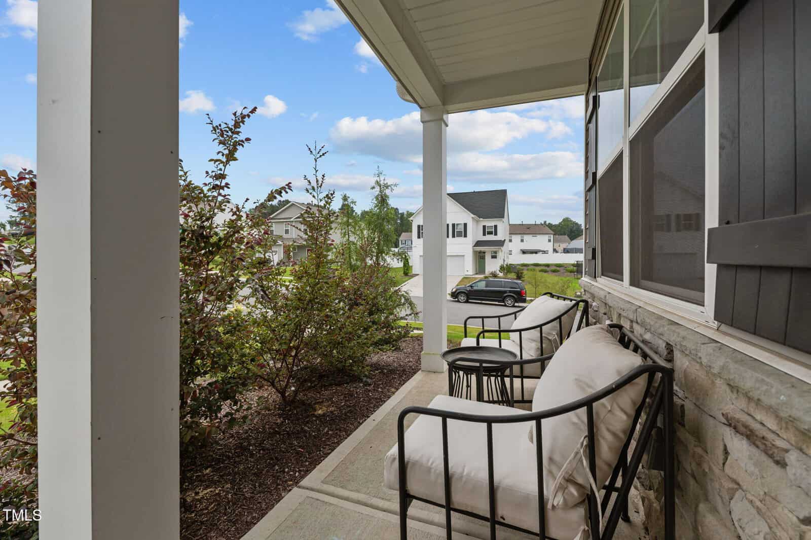 Two cushioned chairs sit on a covered front porch with columns, facing landscaped bushes and a neighborhood street with houses and parked cars visible on a clear day.