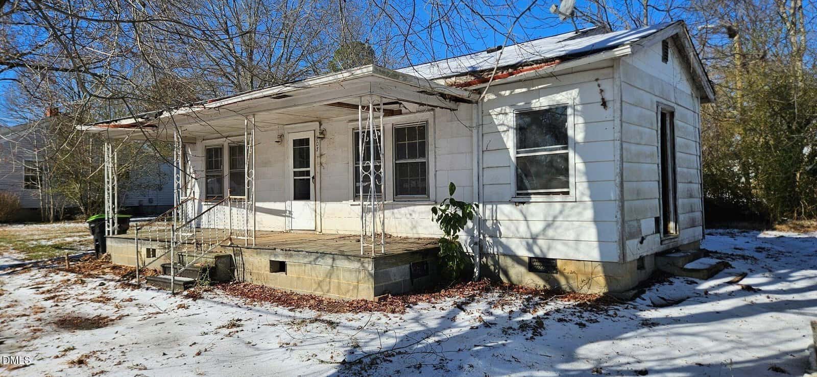 A small, weathered white house with a rusted metal porch roof, metal railings, and a concrete front porch sits on a snowy yard under clear blue skies. The house has peeling paint and overgrown vegetation nearby.