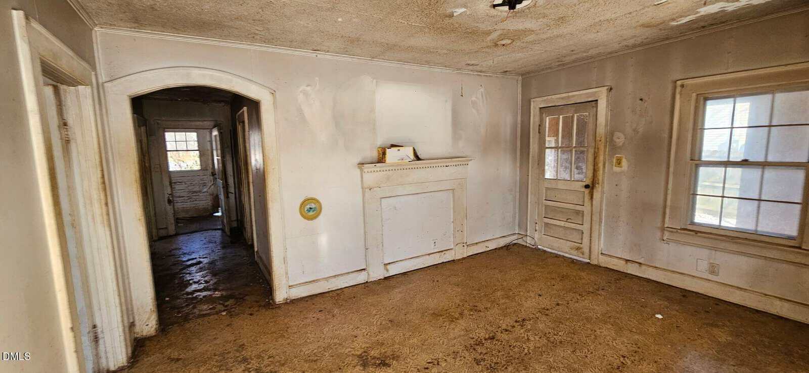 An empty, worn room with peeling paint, dirty floors, a white fireplace, a door with glass panes, two windows, and an open doorway leading to a dimly lit, cluttered hallway.