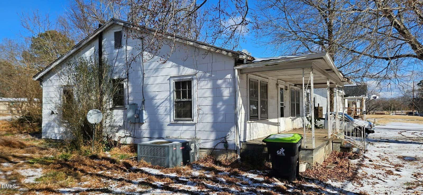 A small, white, single-story house with a covered front porch and several windows. Leafless trees surround the yard, with patches of snow on the ground and porch. A green garbage bin and an air conditioning unit are visible outside.